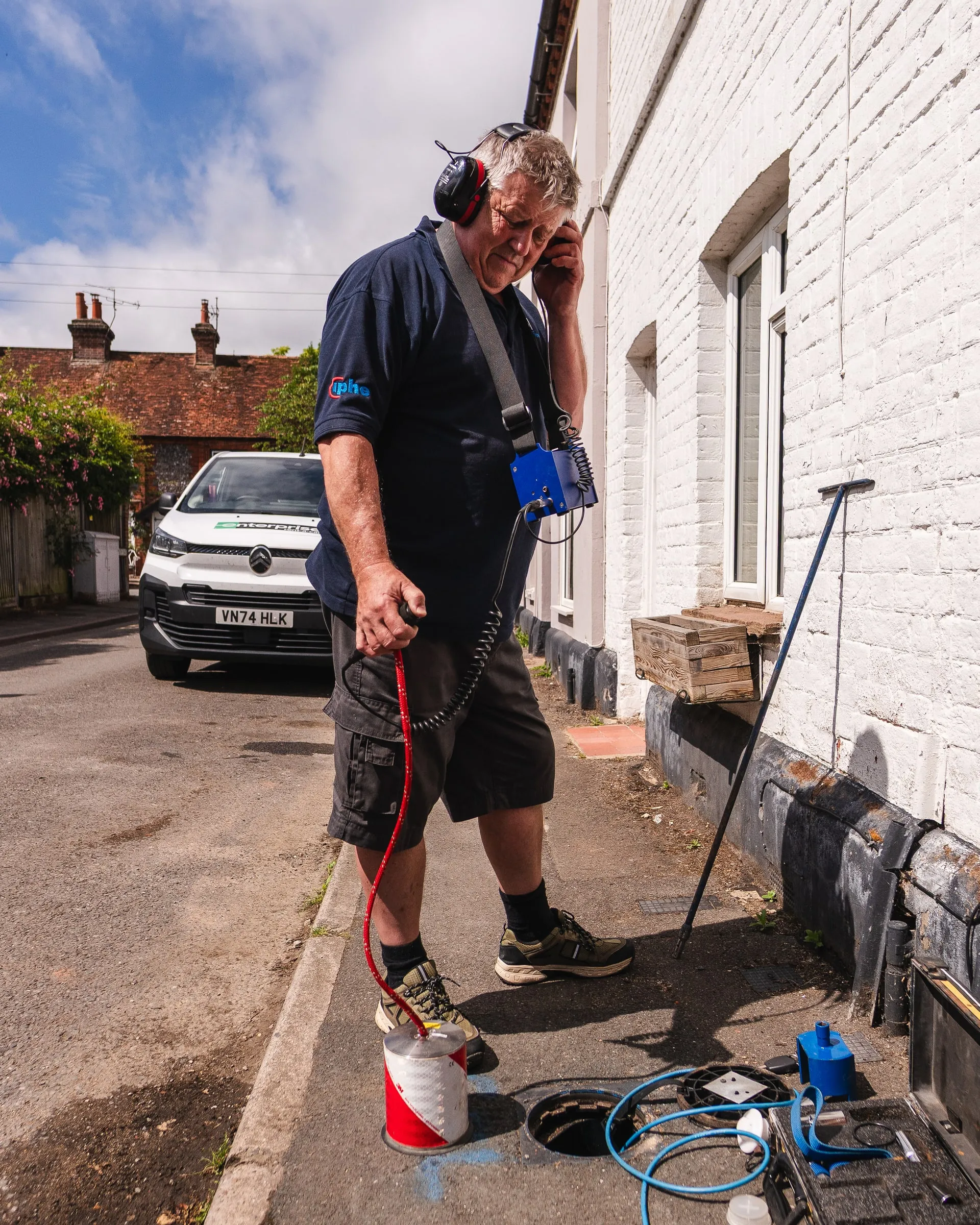 Engineer wearing ear defenders holding a ground-listening probe on a paved access road beside a building, with equipment at his feet