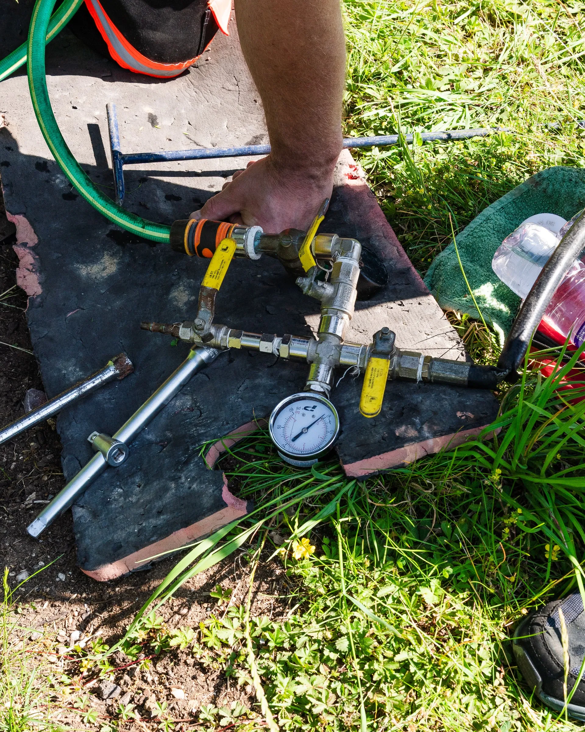 Pressure test rig with valves, hose connections and a gauge laid out on the ground beside a pipe